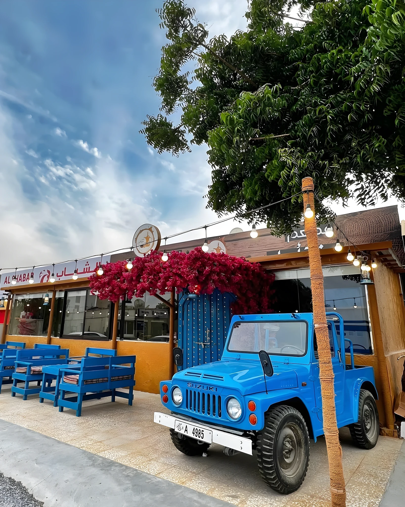A bright blue vintage Suzuki jeep with a UAE license plate parked outside a cafe named Al Shabab, with blue wooden benches, red bougainvillea flowers, string lights, and a green tree overhead against a partly cloudy sky.