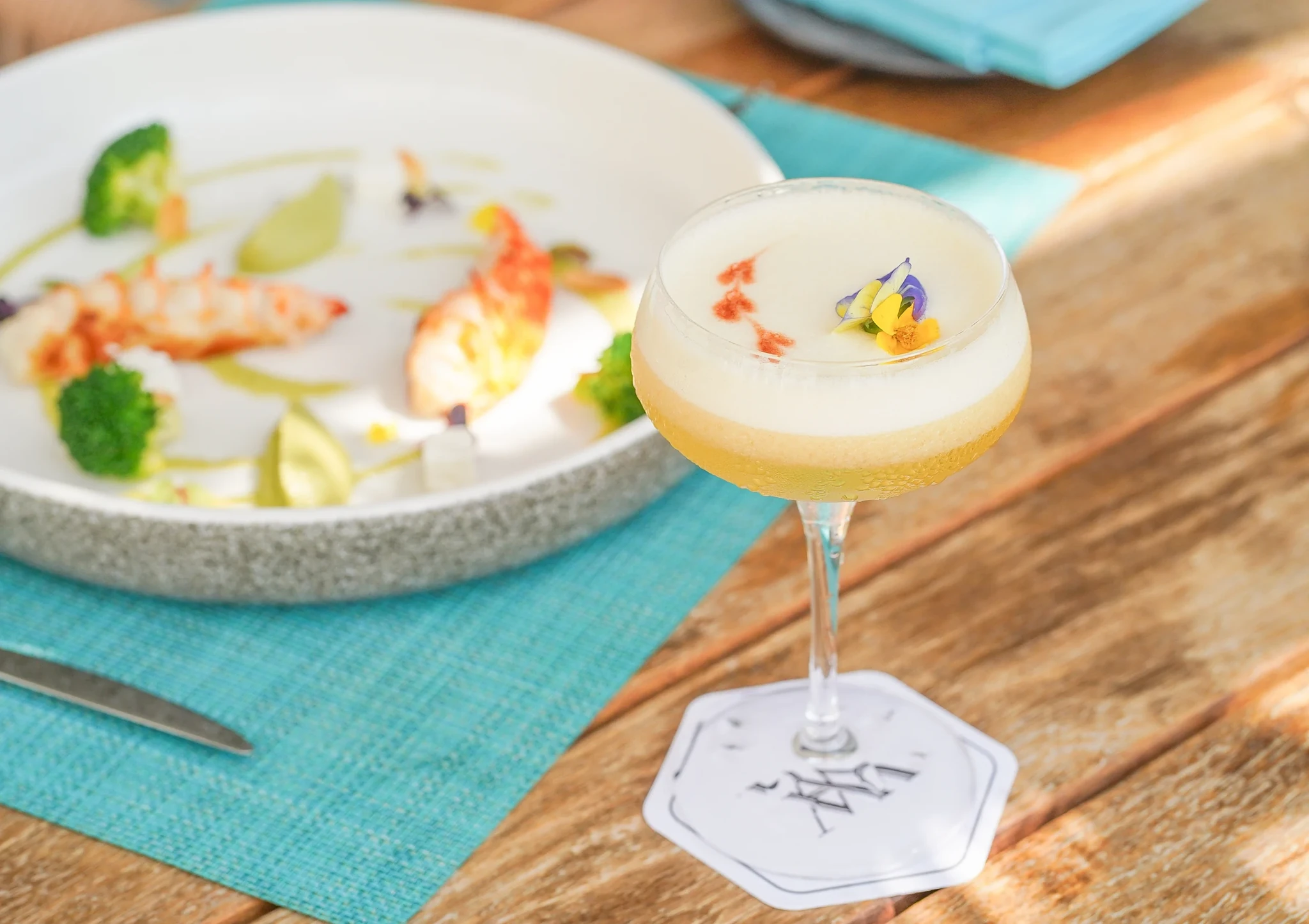 Palm Shore Pool Bar, Ras Al Khaimah: A coupe glass holding a foamy layered cocktail garnished with purple and yellow edible flowers, resting on a white coaster, next to a white bowl plated with prawns, broccoli, and green sauce dots on a turquoise