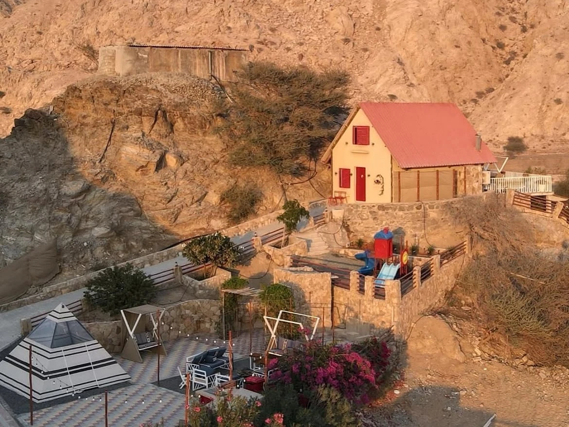 A cream cottage with red roof and red door on a stone terrace built into a rocky mountainside, with a children's playground slide, a pyramid tent, outdoor seating, and pink bougainvillea flowers, beneath a pastel pink and purple sunset sky.