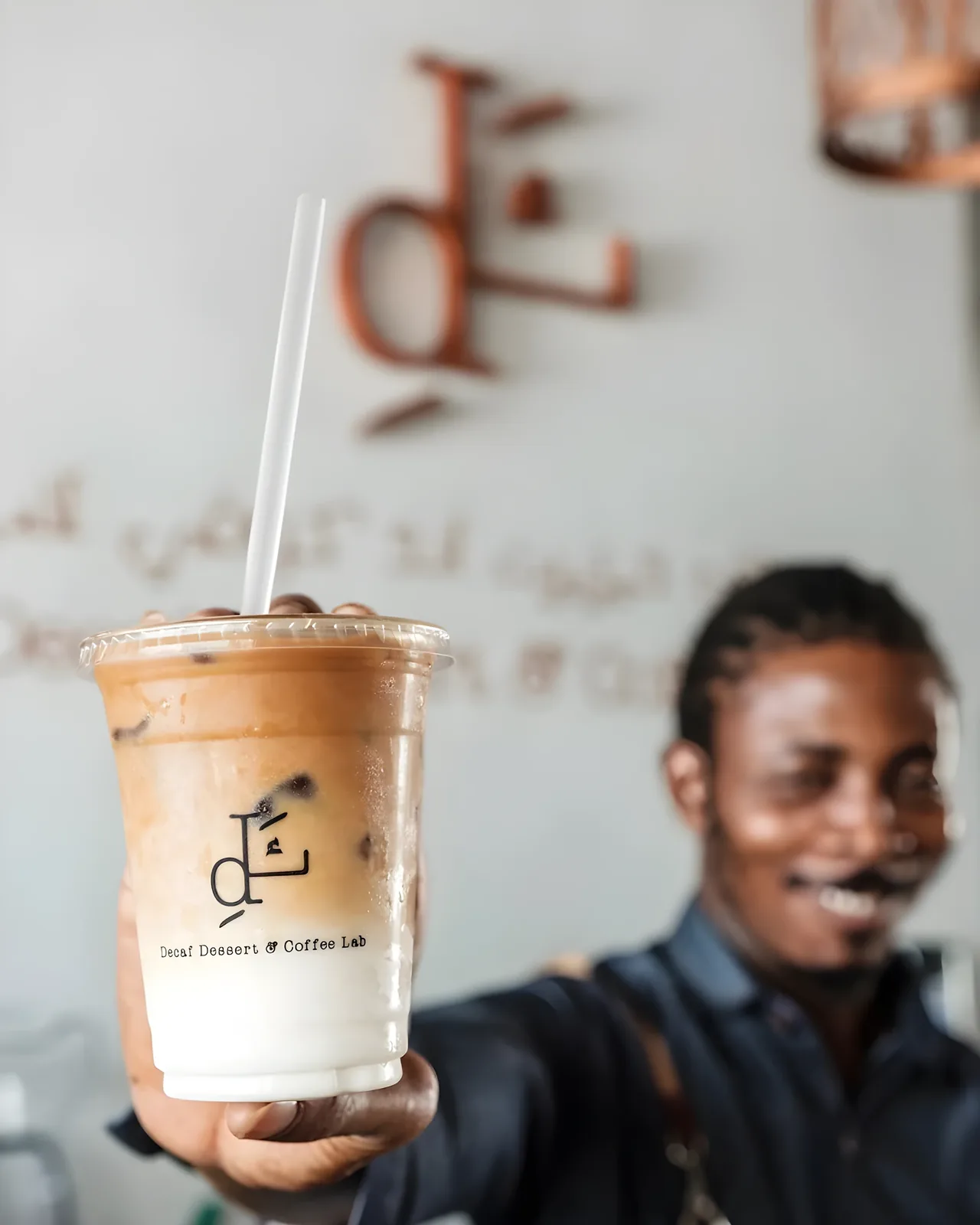 A hand holds a clear plastic cup with a white straw, filled with a layered iced coffee drink over ice, labeled Decaf Dessert & Coffee Lab; a smiling man in a dark shirt and apron stands blurred in the background against a wall with the cafe's logo.