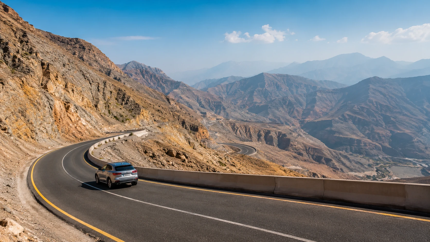 A silver SUV on a curving paved mountain road with yellow lane markings and concrete barrier, surrounded by rocky brown peaks and distant hazy mountains under a clear blue sky.
