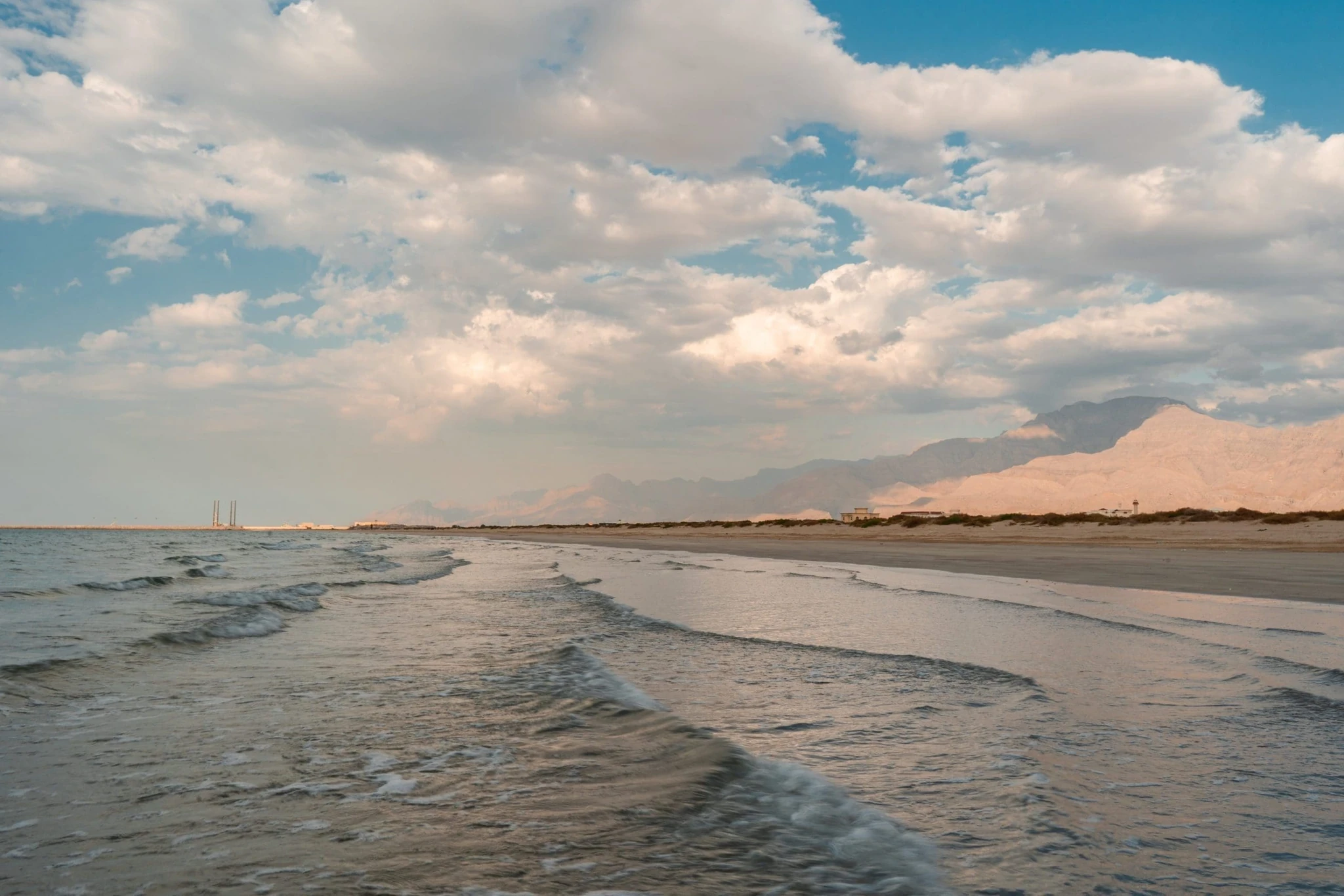 A tranquil beach with waves breaking on wet sand, a flat shoreline with low buildings and towers in the distance, and rugged mountains beneath a bright sky filled with soft clouds during the late afternoon.