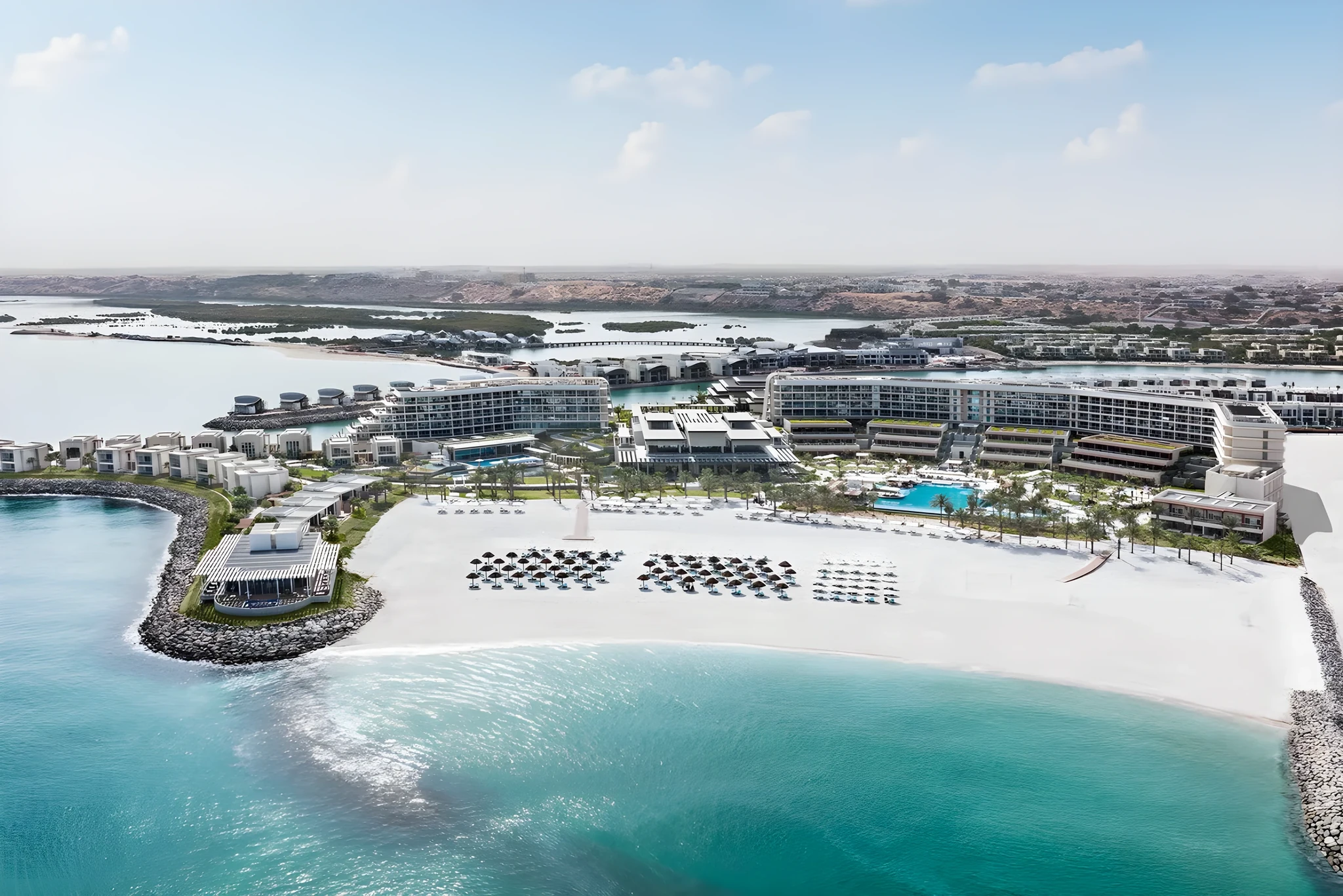 Aerial photograph of a coastal resort featuring curved low-rise white buildings, a large swimming pool lined with palm trees, a white-sand beach with organized rows of parasols and sun loungers, and clear turquoise sea in the foreground under a pale blue sky.