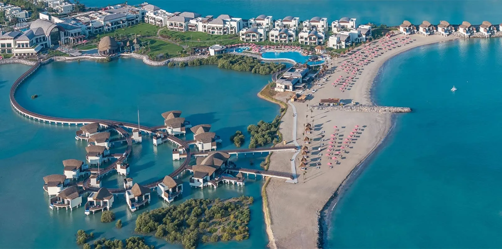 Aerial photograph of a coastal resort with thatched-roof overwater bungalows linked by wooden walkways over turquoise water, a crescent sandy beach dotted with rows of pink sun umbrellas and loungers, a lagoon-style pool, and clusters of white low-rise villa buildings surrounded by greenery.