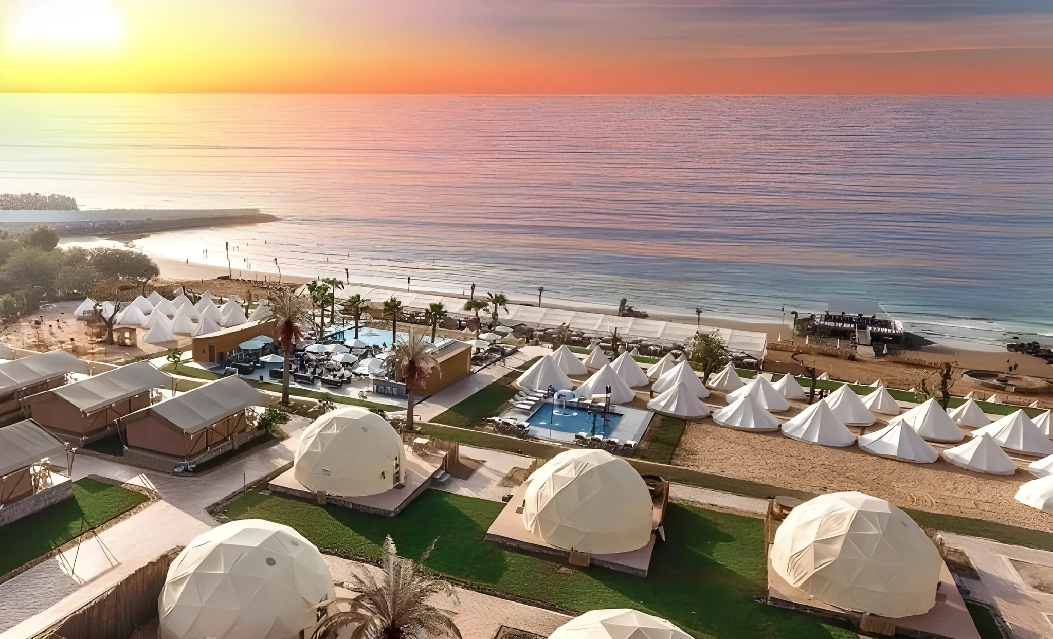 Aerial view of a beachfront glamping resort with white geodesic dome tents and pointed bell tents arranged on green lawns beside pools and palm trees, facing a sandy beach and calm sea under an orange sunset sky.