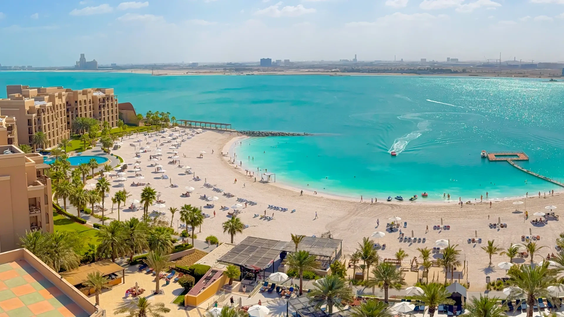 Aerial view of a beige beachfront resort with palm trees, white parasols and sun loungers lining a wide sandy beach that curves along a clear turquoise bay, with swimmers, jet skis and a boat in the water and a hazy coastline in the distance.