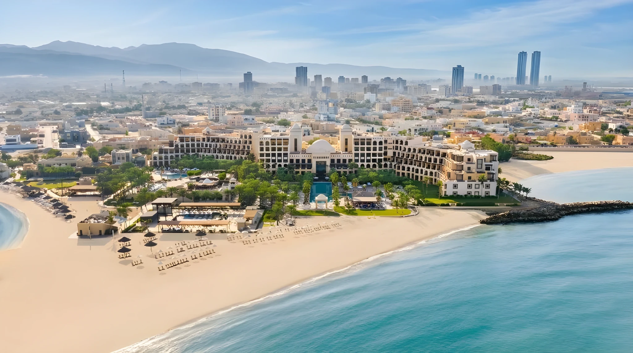 Aerial view of a large domed beachfront resort surrounded by palm gardens and pools, fronted by a wide sandy beach with rows of umbrellas and loungers, calm turquoise sea, a stone breakwater, and a city skyline with high-rise towers and mountains in the hazy background.