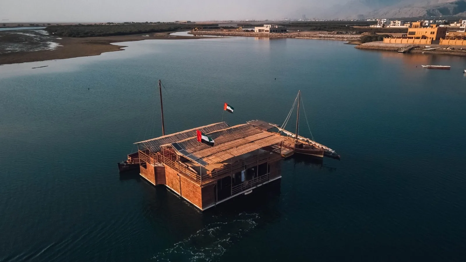 Aerial view of a large wooden floating lodge with palm-thatch roof and two UAE flags on a calm blue-green lagoon, with a moored dhow, mangroves, buildings, and mountains in the background.