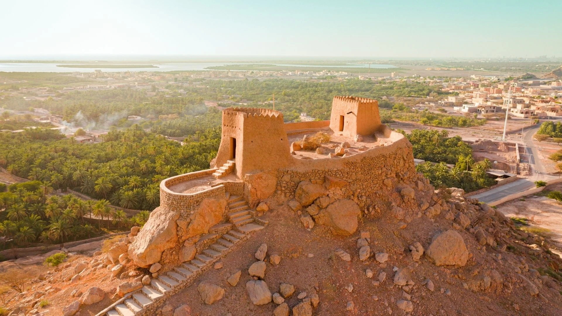 Aerial view of an old sand-colored stone and mud-brick fort with crenelated towers atop a rocky hill, stone steps leading up, overlooking green palm plantations, a town, and a coastal lagoon under a hazy sky.