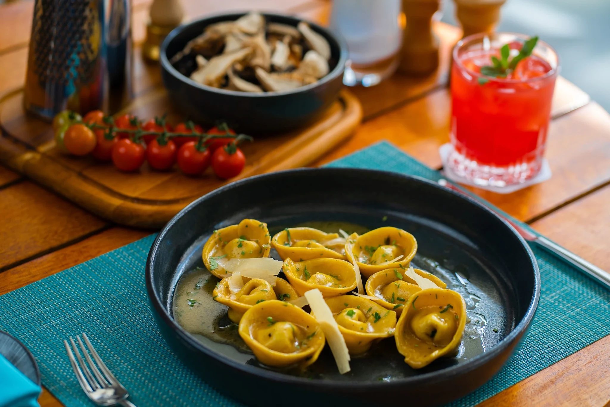 Azure, Ras Al Khaimah: Black plate of golden tortellini in butter sauce topped with shaved parmesan and chopped parsley, set on a teal woven placemat over a wooden table, with a dark bowl of sliced mushrooms, cherry tomatoes on the vine, and a red