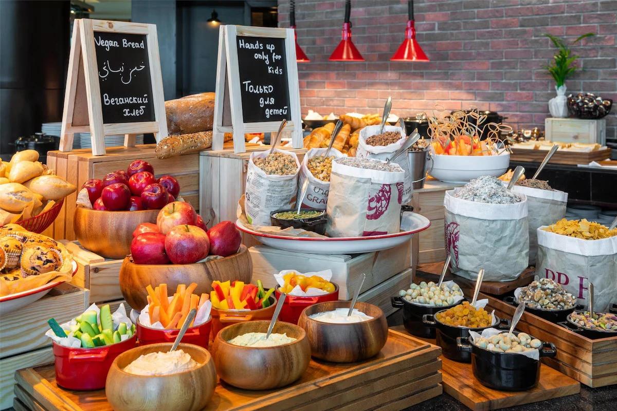 Buffet counter with wooden bowls of red apples, cut vegetable sticks, dips, paper sacks of granola and cereal, breads, and pastries under red hanging heat lamps, with chalkboard signs reading Vegan Bread in several languages.