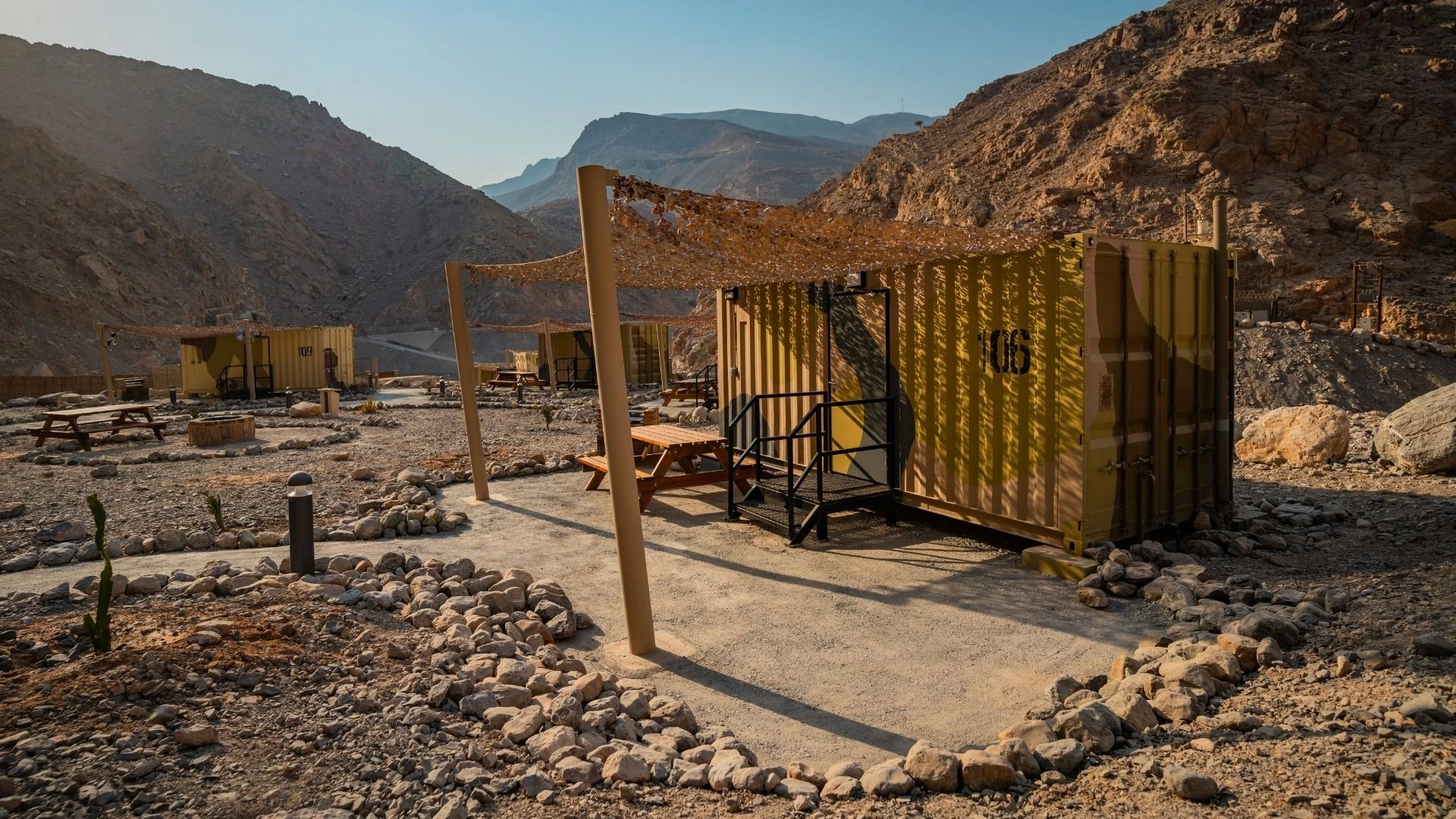 Camouflage-painted shipping container cabins with metal steps, wooden picnic tables and camo shade netting set on gravel amid rocky brown mountains at golden hour.