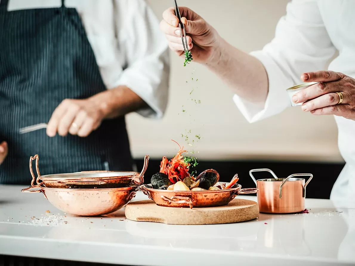 Chef using tongs to garnish a copper serving dish of langoustine, mussels and clams with chopped herbs, set on a cork board beside copper cookware on a white counter.