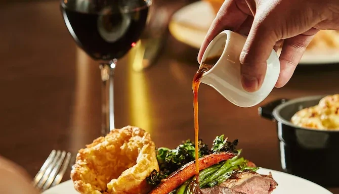 Long Weekend in Ras Al Khaimah: What's On and What It Costs, Ras Al Khaimah: Close-up of gravy being poured from a white jug over a roast dinner plate with Yorkshire pudding, roasted carrot, broccolini, and sliced meat, with a glass of a red drink