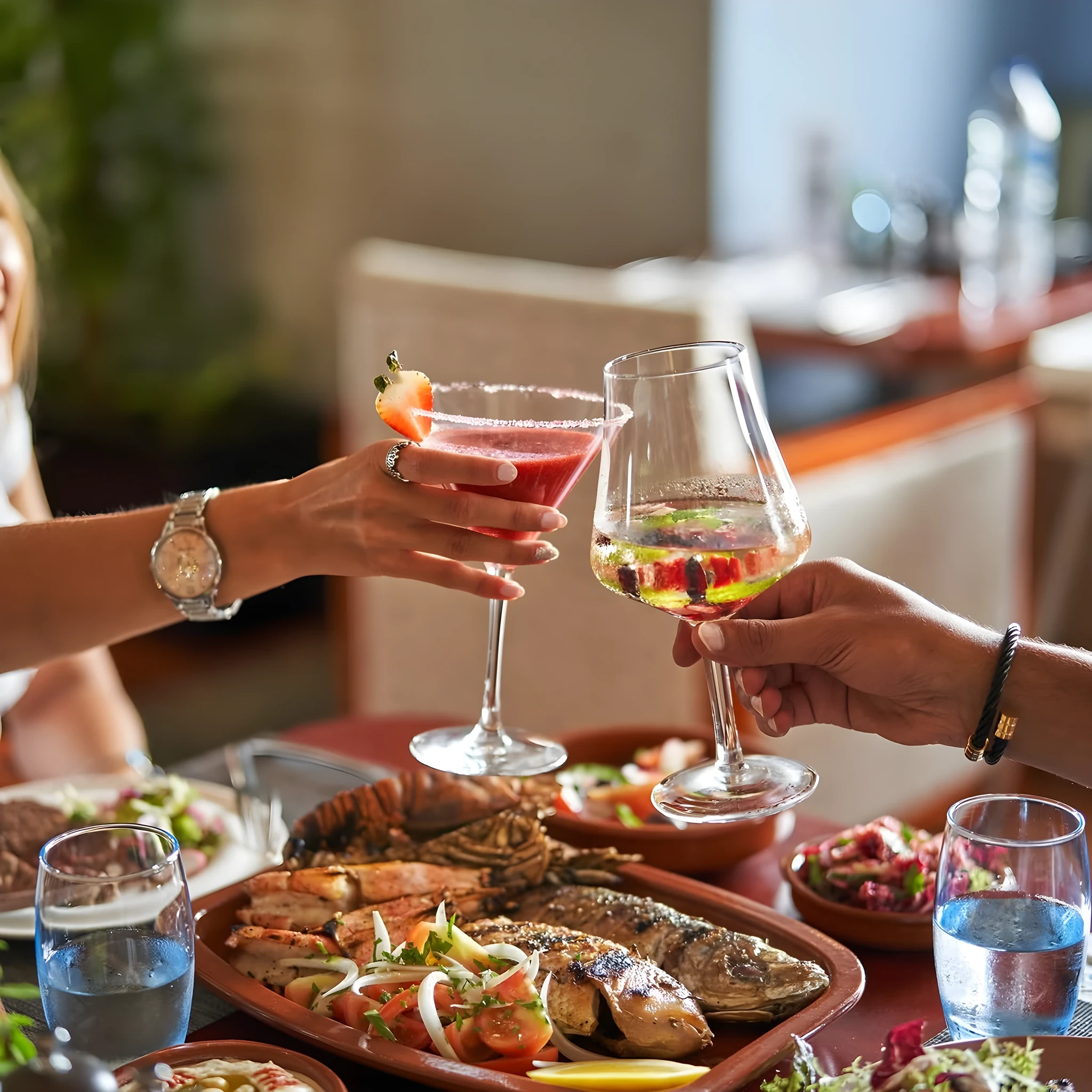 Close-up of two hands toasting a pink strawberry cocktail and a glass of white wine above a platter of grilled lobster, fish and prawns with tomato salad, water glasses and side dishes on a restaurant table.