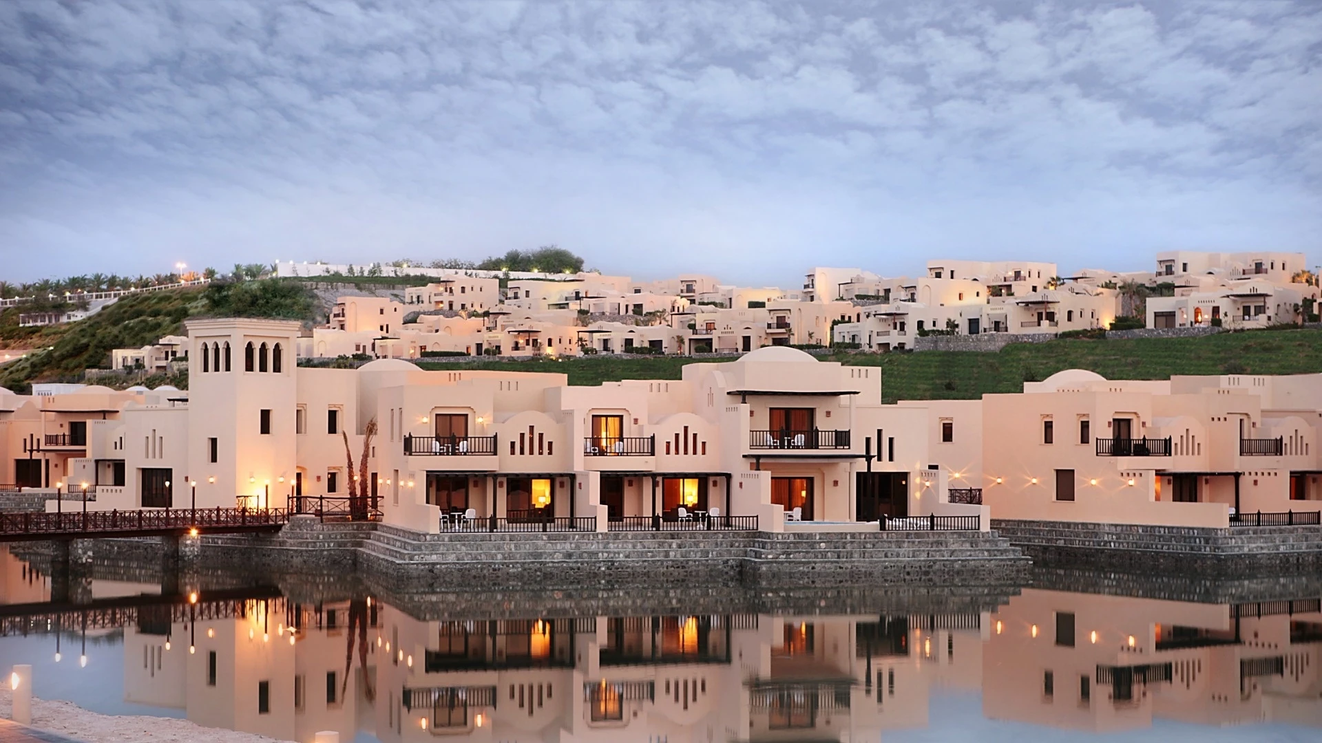 Cream-colored Arabesque villas with domes, arched balconies and warm interior lighting rise along a green hillside, reflected in still water under a cloudy blue evening sky.