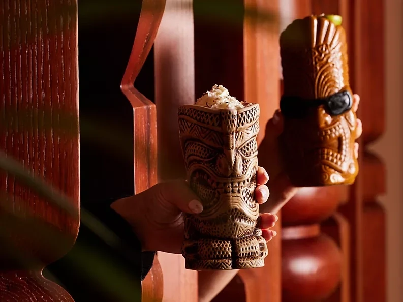 Long Weekend in Ras Al Khaimah: What's On and What It Costs, Ras Al Khaimah: Two hands holding intricately carved tiki mugs, one topped with a foamy garnish, against a backdrop of red-toned wood in warm lighting.