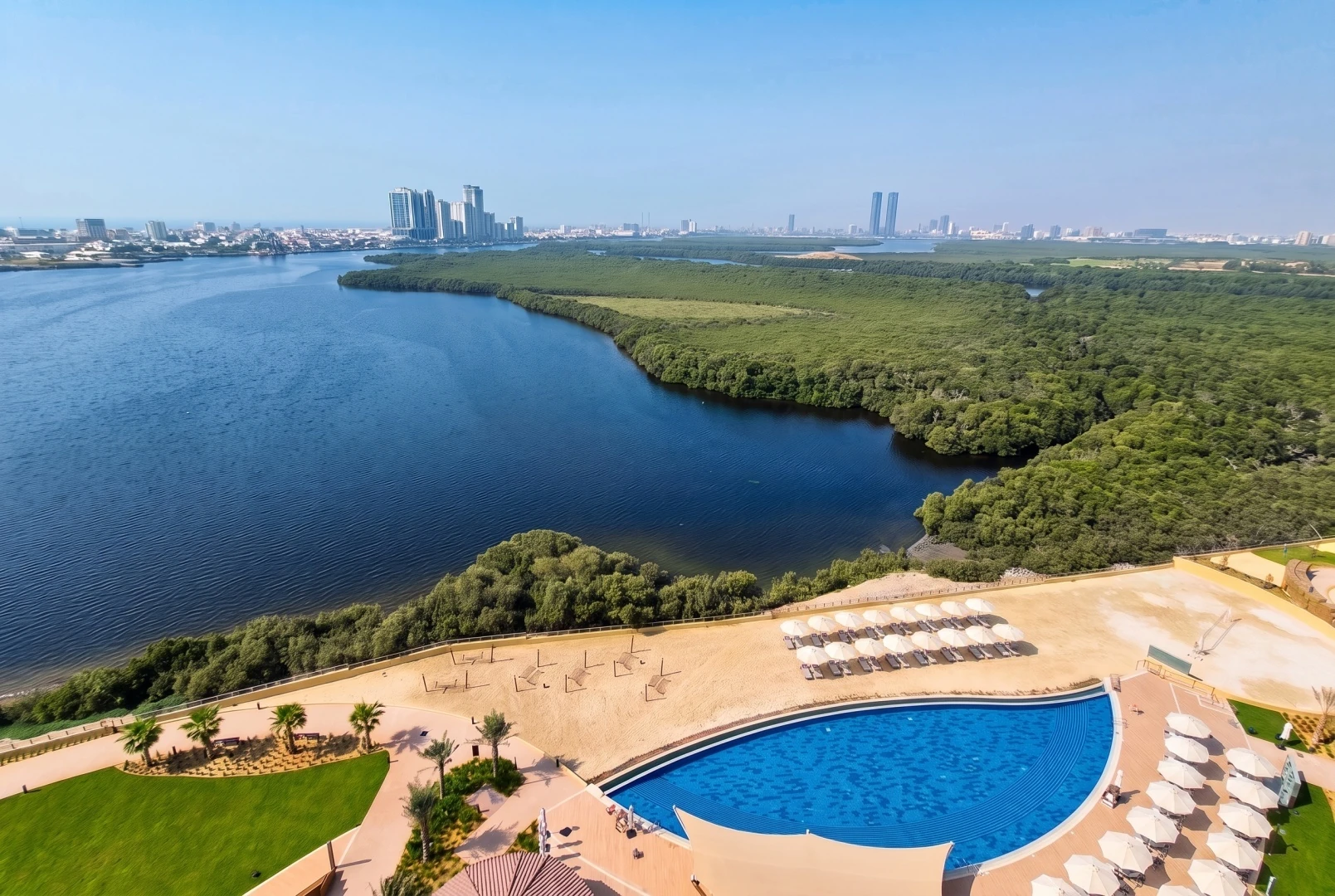 Elevated view of a resort with a large curved blue swimming pool, sandy beach dotted with white umbrellas and sun loungers, green lawns and palm trees, a wide creek bordered by dense green mangroves, and a hazy high-rise skyline on the horizon.