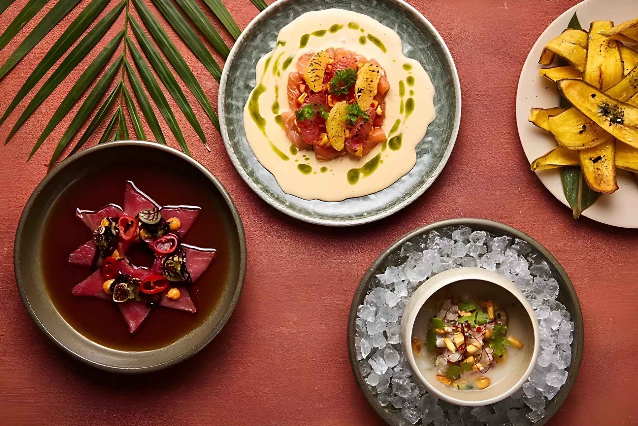 Flat lay of four dishes on a terracotta surface: sliced tuna in dark broth with chili and corn, a citrus-topped ceviche in green sauce, a bowl of ceviche set in crushed ice, and a plate of fried plantain chips, with a green palm leaf.