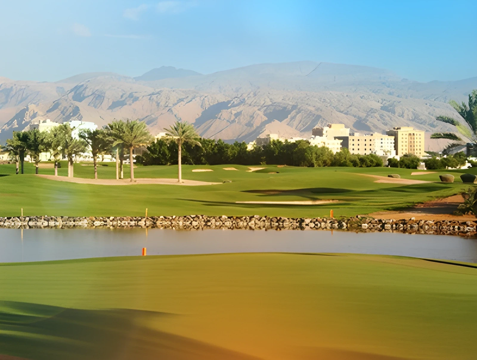Green golf course with a water feature in the foreground, palm trees and low resort buildings in the midground, and rocky mountains rising in the background under a pale blue sky.