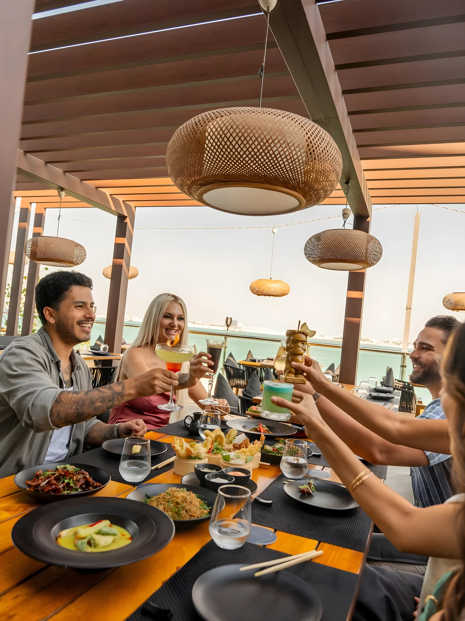 Group of four people toasting colorful cocktails over a wooden dining table filled with black plates of Asian food, seated under woven rattan pendant lights on an open-air terrace overlooking calm blue water.