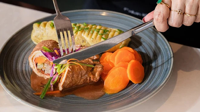 Hands holding a fork and knife cutting a meat dish plated with sliced carrots, potatoes, red cabbage garnish, and brown gravy on a blue plate.