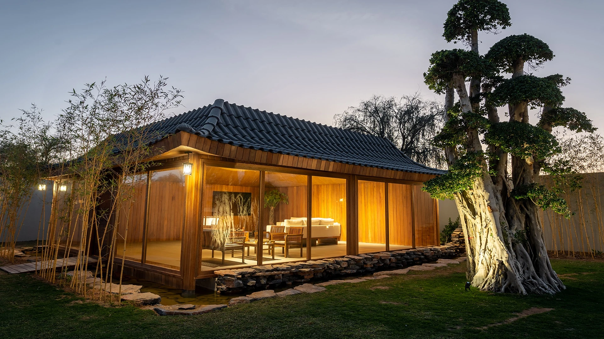 Illuminated wooden pavilion with dark tiled curved roof and glass walls showing lounge seating inside, set among bamboo, a stone-lined water pond, and a large manicured tree on a lawn at dusk.