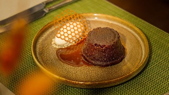 Individual sticky toffee pudding cake in caramel sauce, topped nearby with cream and a honeycomb-patterned sugar tuile, served on a green and gold ceramic plate over a woven green placemat with cutlery in the background.