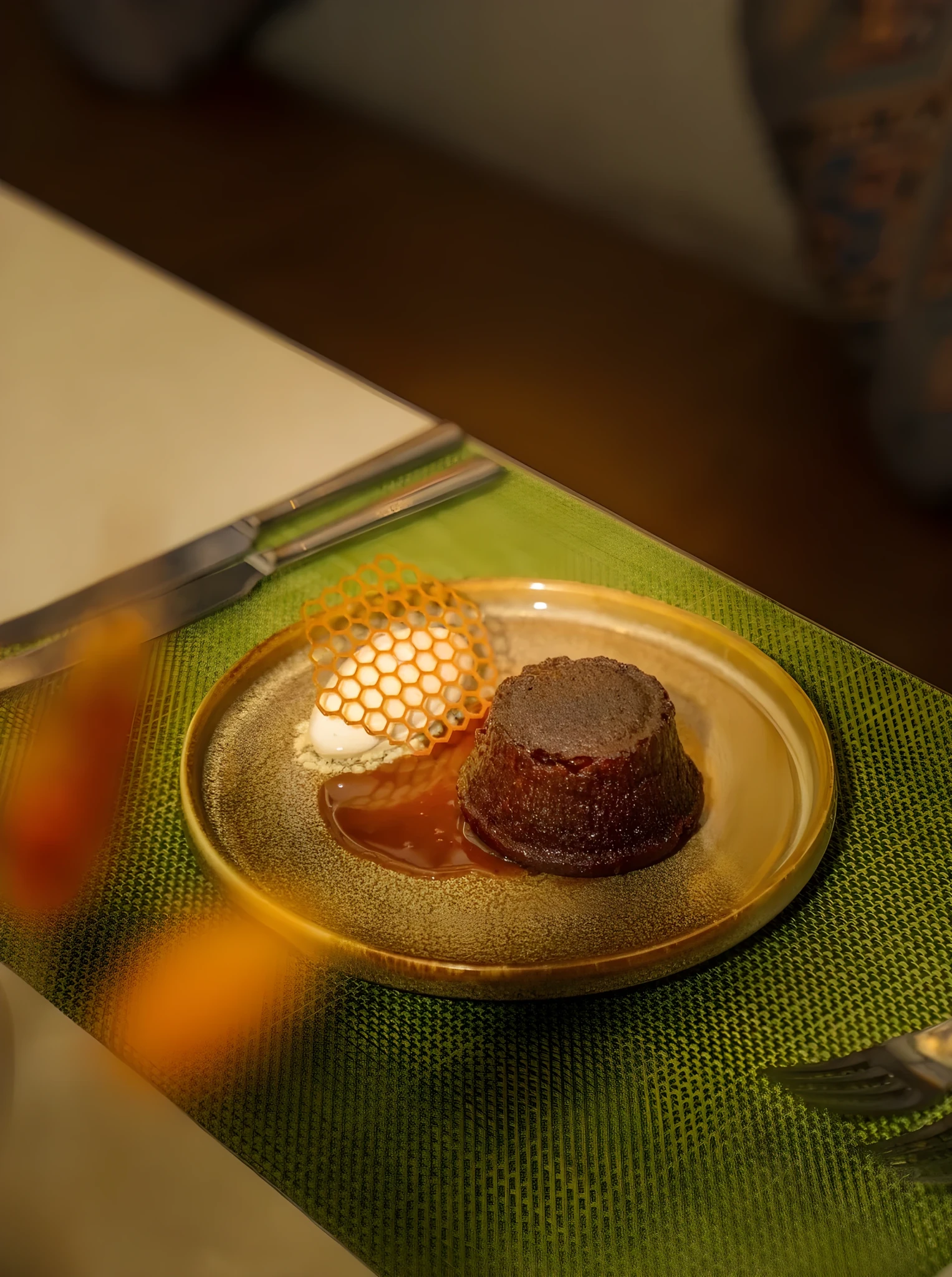 Individual sticky toffee pudding with caramel sauce and a scoop of cream, topped with a honeycomb-patterned caramel tuile, plated on a speckled brown-and-gold ceramic dish set on a green woven placemat with a knife nearby.