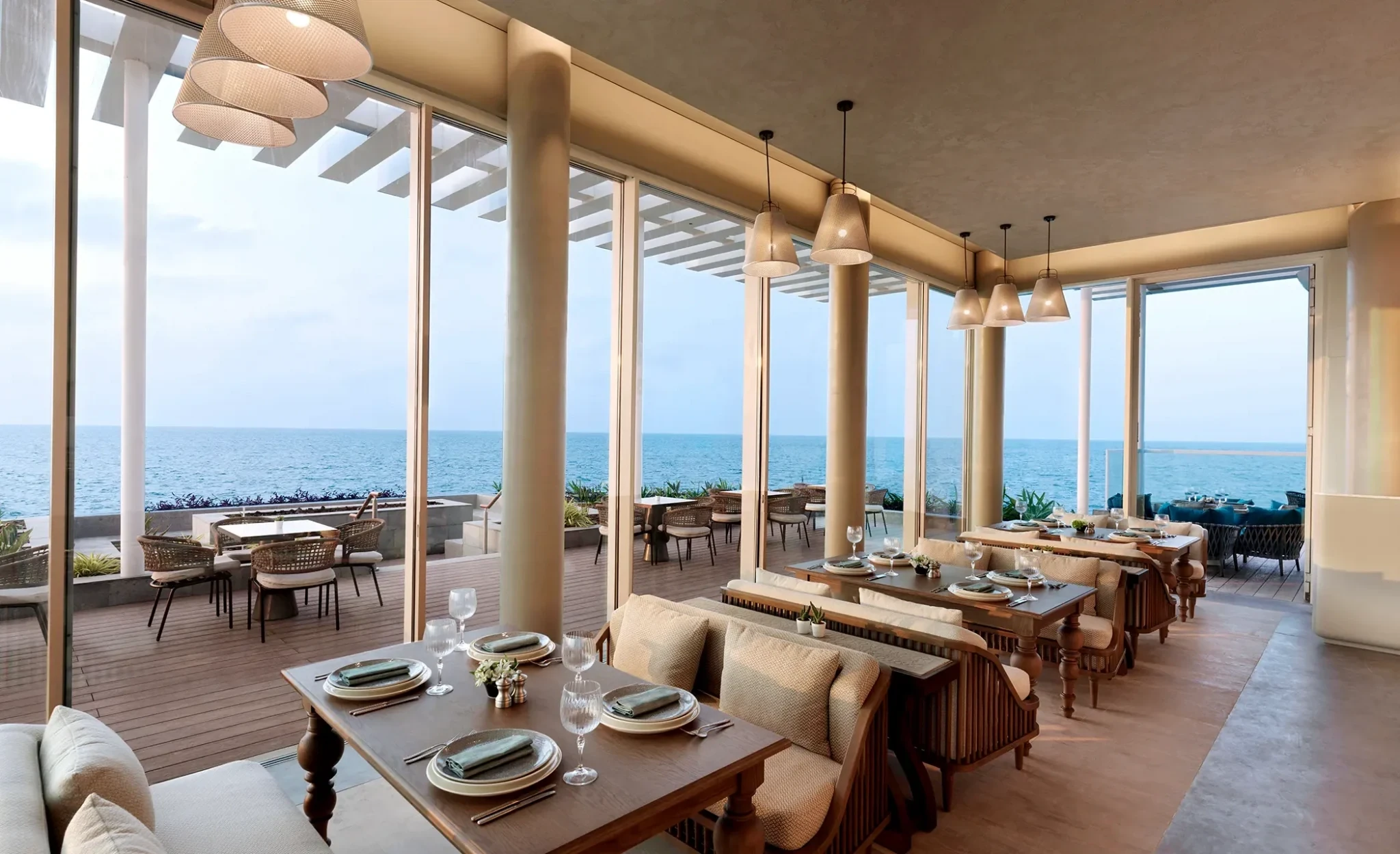 Interior of a coastal restaurant featuring wooden dining tables set with plates, napkins and wine glasses, cushioned bench seating, woven pendant lamps, and large glass windows revealing an outdoor terrace and the ocean beyond.