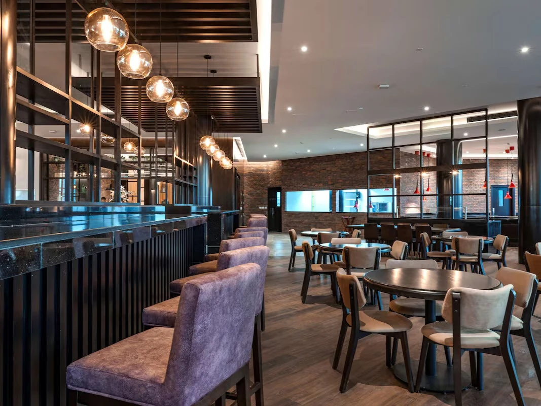 Interior of a modern hotel bar and lounge with a long black stone counter, purple velvet bar stools, spherical glass pendant lights, exposed brick walls, and round wooden tables with cushioned chairs on a wood floor.
