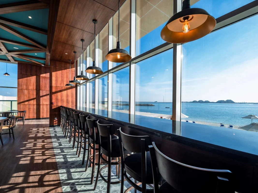 Interior of a waterfront bar with black high stools along a wooden counter, industrial pendant lights, wood-paneled walls, and large windows showing a blue ocean, sandy beach, and parasols.