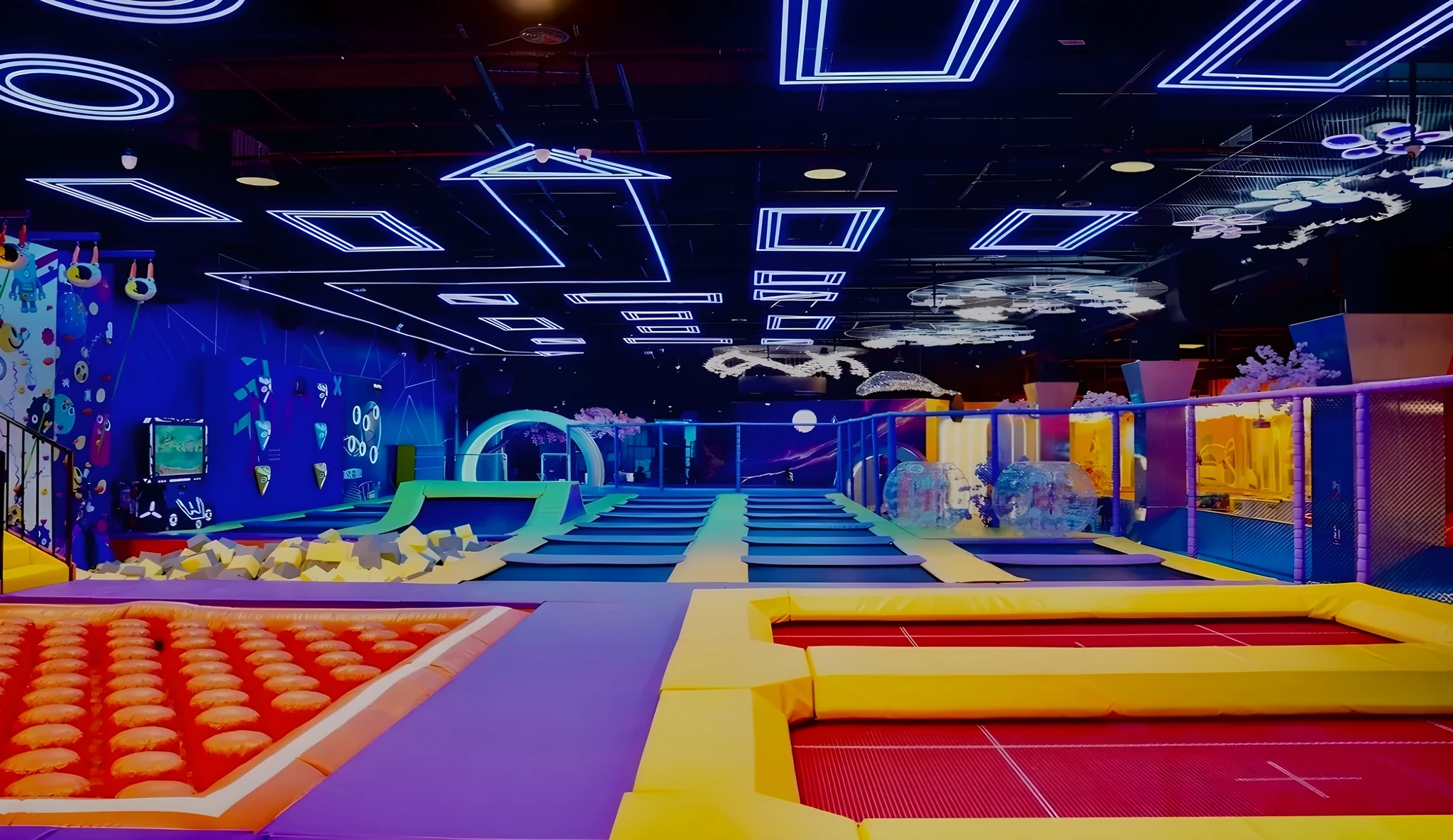 Interior of an indoor trampoline park with purple, yellow, and blue padded jumping areas, an orange dome trampoline section, a foam pit, a climbing wall, and blue neon geometric lights on a dark ceiling.