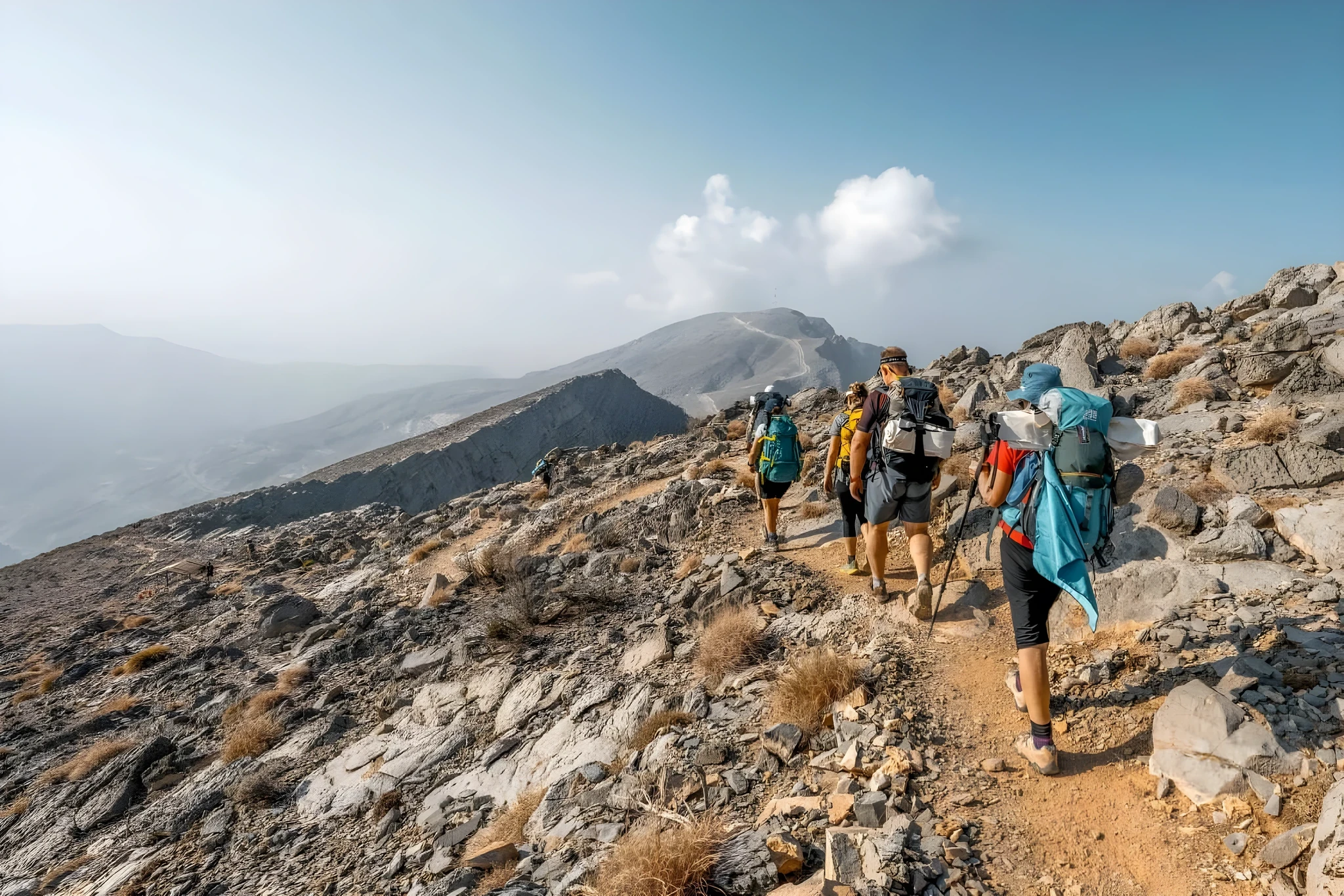 Line of hikers carrying large backpacks and trekking poles ascending a rocky dirt trail along a mountain ridge, with hazy peaks and a bright cloud in the blue sky in the distance.