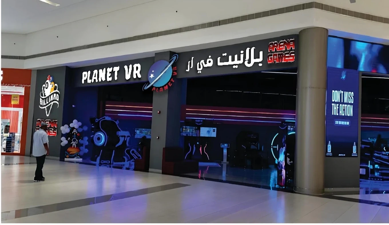Mall storefront of Planet VR arcade featuring illuminated planet logo, English and Arabic signage, neon-lit gaming interior, and a person walking across the tiled floor.