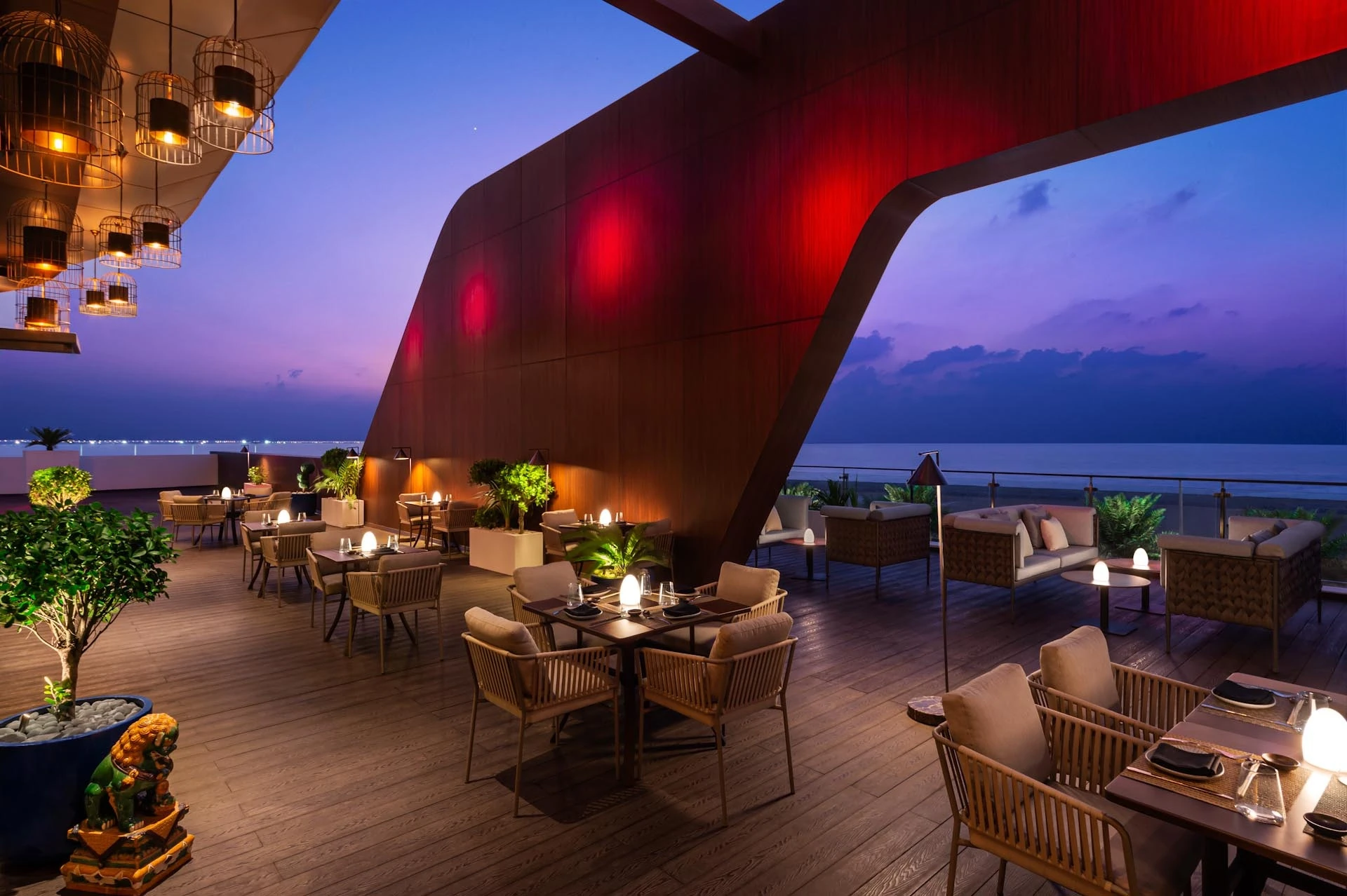 Outdoor restaurant terrace with wooden decking, set dining tables and cushioned chairs, birdcage-style pendant lights, a curved red-lit wood wall, and lounge sofas overlooking the sea at dusk under a purple sky.