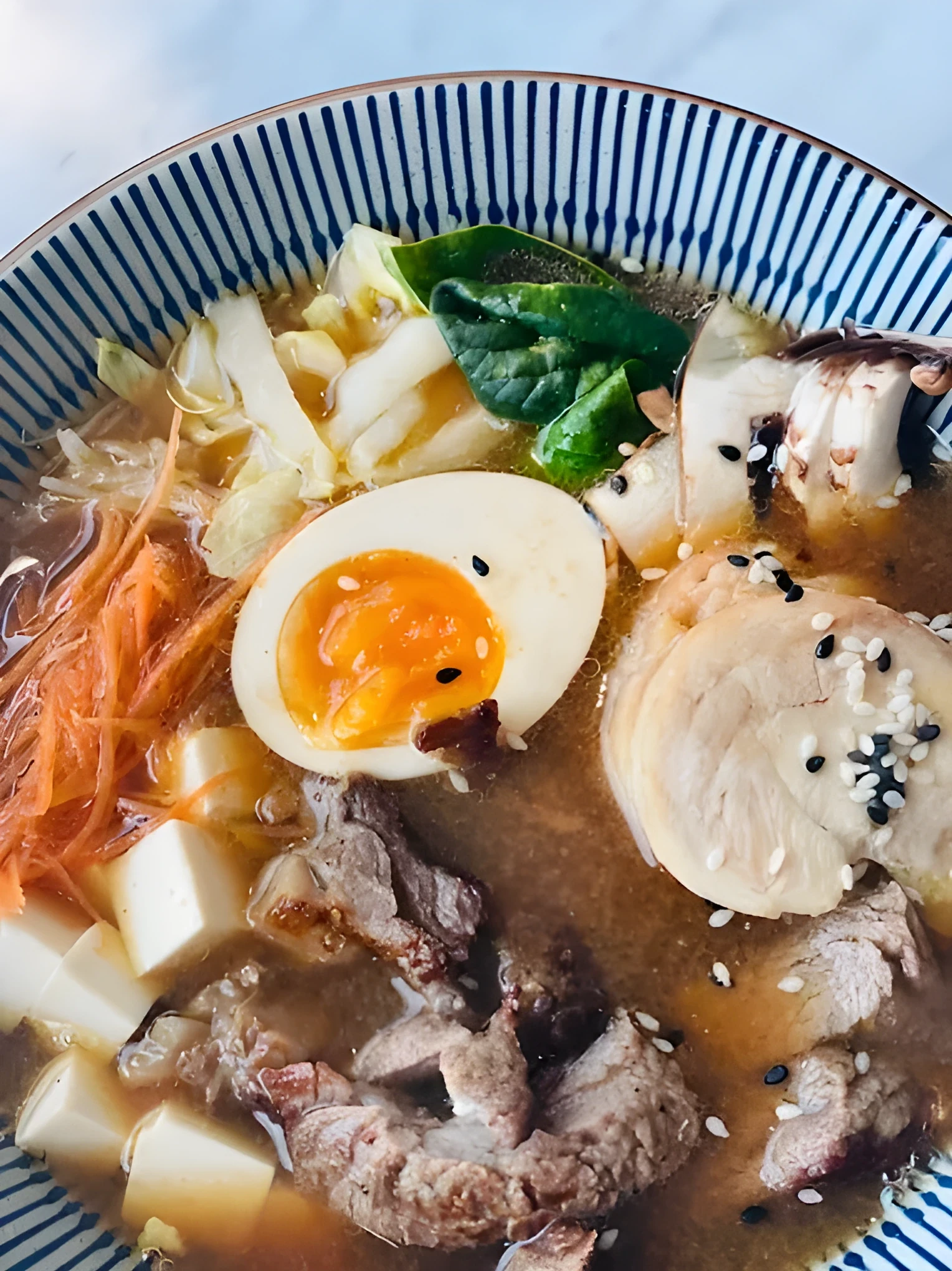 Overhead close-up of a ramen bowl with blue and white patterned rim, holding a halved soft-boiled egg with runny yolk, slices of beef, cubed tofu, shredded carrot, cabbage, spinach, and sesame seeds in brown broth.