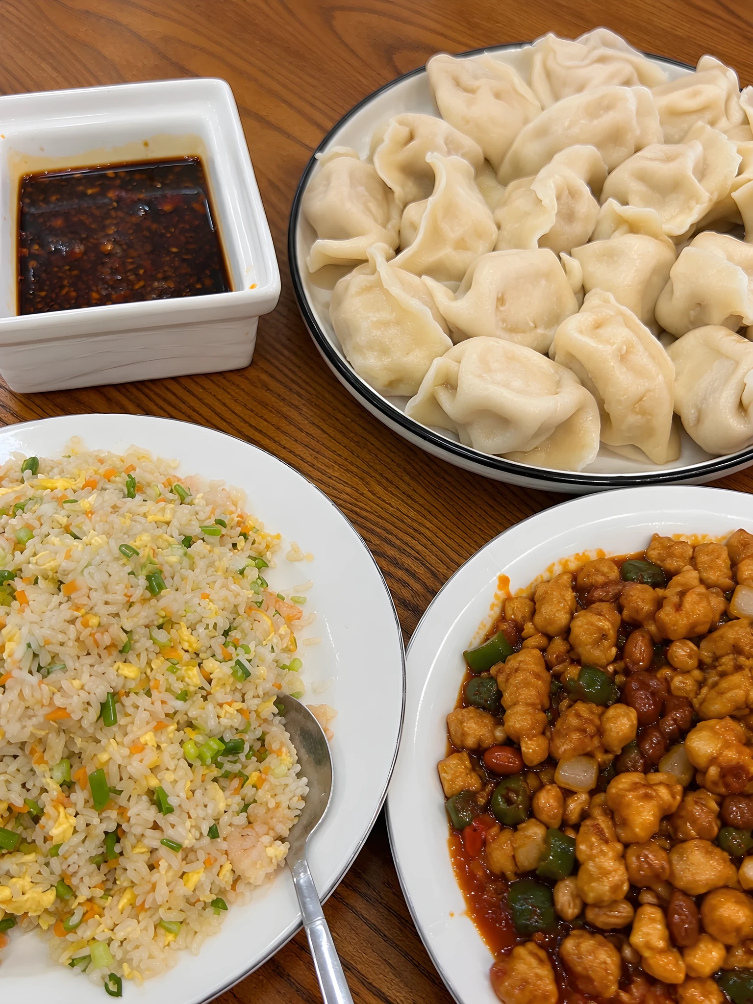 Overhead view of a Chinese meal on a wooden table featuring a plate of boiled dumplings, a plate of egg and vegetable fried rice with a spoon, a plate of spicy diced chicken with peanuts and green peppers, and a white square dish of dark chili dipping sauce.