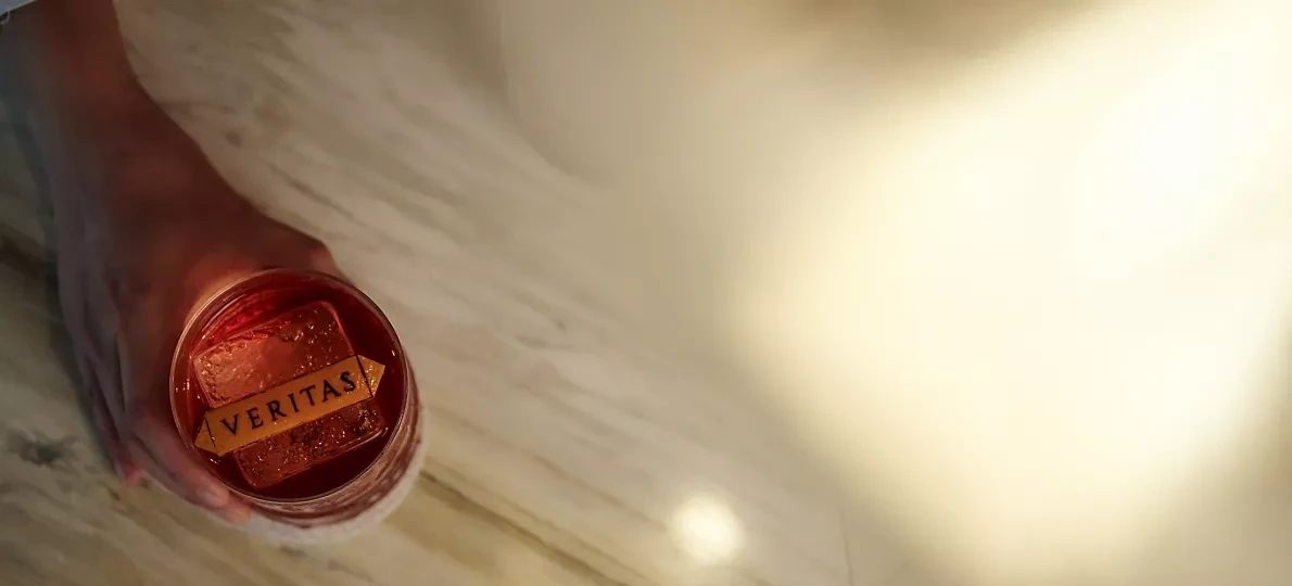 Overhead view of a red cocktail in a glass with a clear ice cube branded Veritas in gold lettering, held over a light marble bar top with warm lighting.