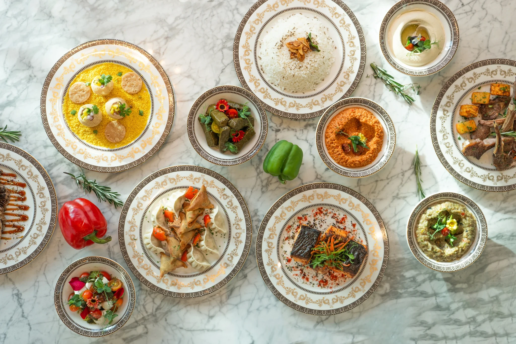 Qasr al Bahar, Ras Al Khaimah: Overhead view of gold-rimmed plates and bowls on white marble holding steamed rice, samosas, stuffed vine leaves, red and green dips, seared fish, lamb chops, saffron rice, and salads, with fresh rosemary and red and