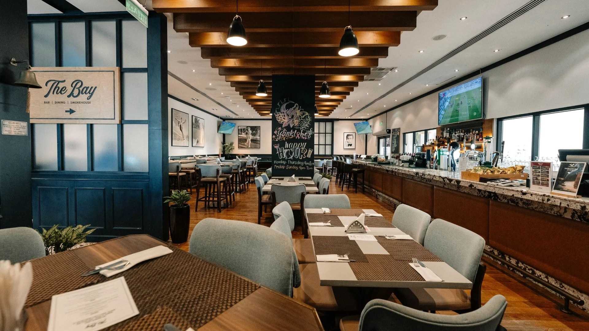 Restaurant and bar interior with wooden tables set with cutlery, grey-green upholstered chairs, a long bar with taps and bottles on the right, a wooden 'The Bay' sign on a navy blue wall, a chalkboard happy hour sign, pendant lights and a wall-mounted TV showing sports.
