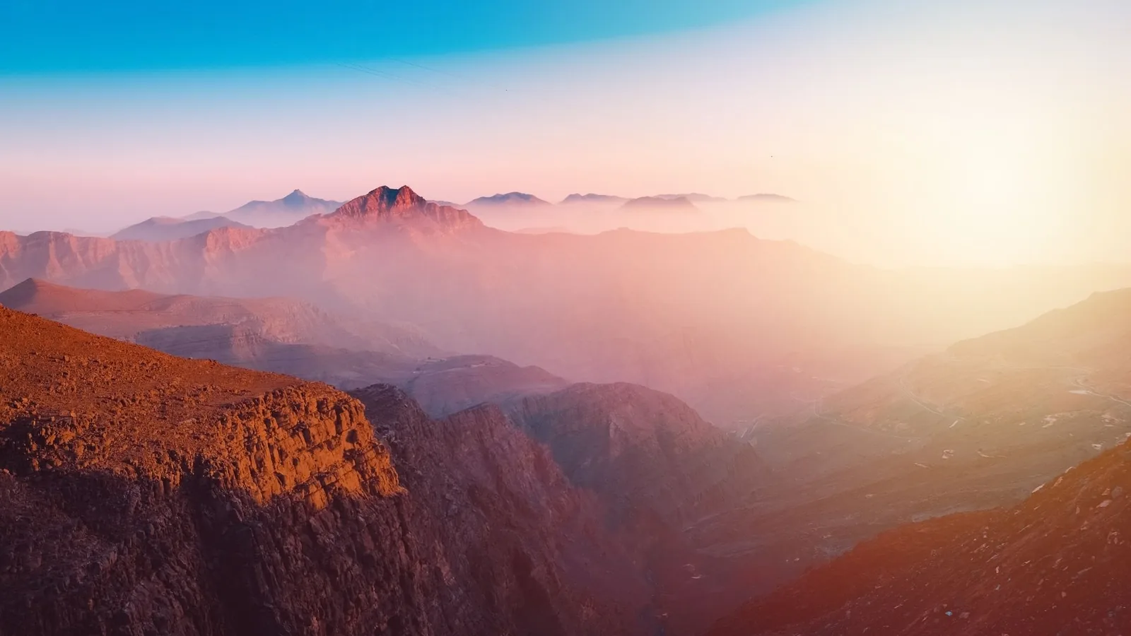 Rocky mountain landscape with jagged peaks and a deep canyon bathed in soft orange and pink haze under a clear blue-to-warm gradient sky at sunrise.