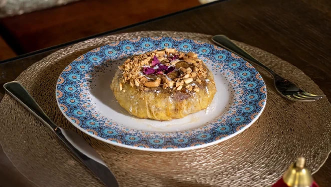 Round golden pastry topped with chopped nuts and a dried rose petal, served on a white plate with a blue and orange mandala border, set on a woven placemat with cutlery on either side.