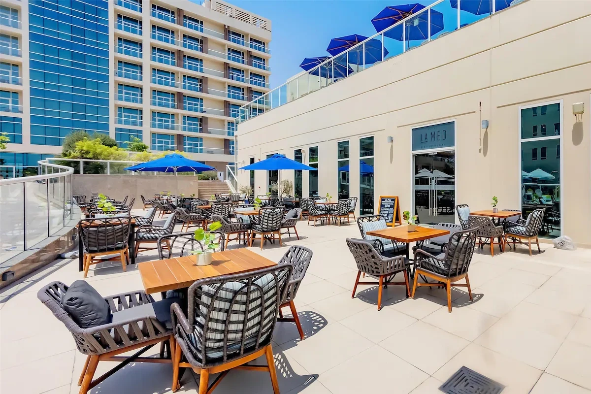 Sunlit outdoor dining terrace with wooden tables, gray woven rope armchairs, blue parasols, and a glass-fronted restaurant entrance labeled La Med, set beside a tall building against a blue sky.