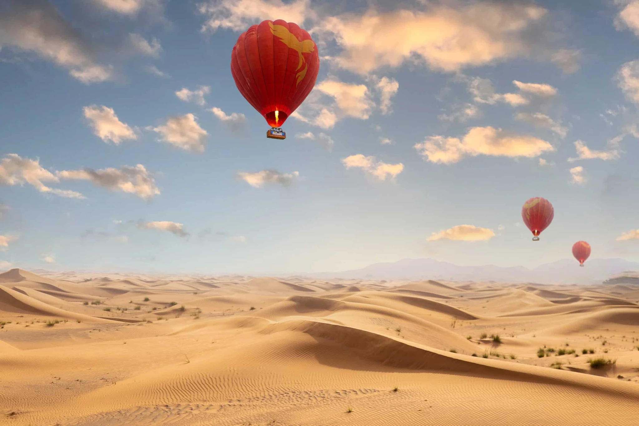 Three red hot air balloons with yellow bird designs floating above rippled desert sand dunes with distant mountains under a partly cloudy blue sky.