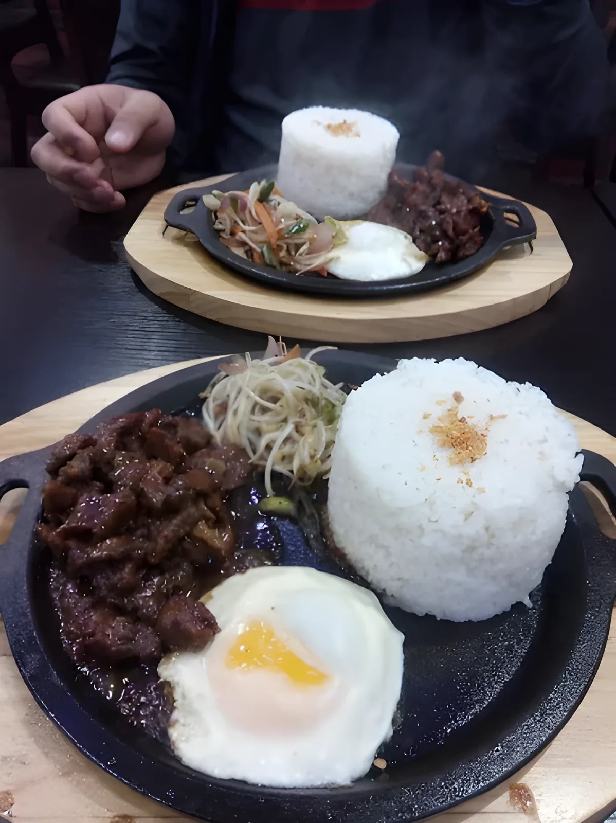 Two black sizzling skillets on wooden boards, each holding marinated beef, a sunny-side-up fried egg, a mound of white rice, and vegetables, with a person seated in the background.