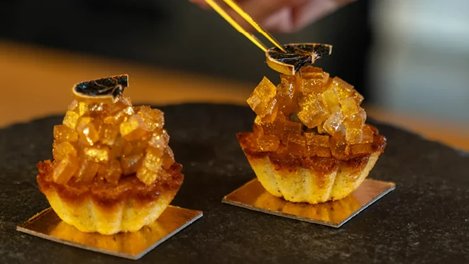 Two individual cakes on gold square boards, each topped with glossy amber candied citrus cubes and a dried orange slice, with a hand using gold tongs to garnish one on a dark stone surface.