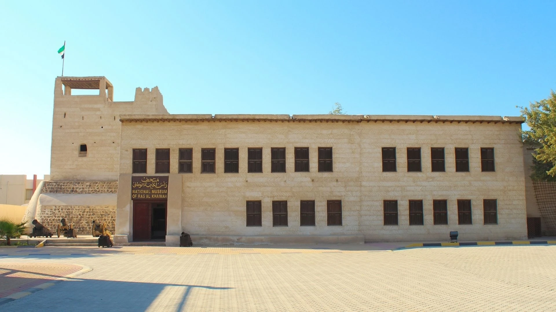 Two-story sand-colored former fort with a corner watchtower flying the UAE flag, dark shuttered windows, a signboard reading 'National Museum of Ras Al Khaimah', and antique cannons on a paved courtyard under a clear blue sky.