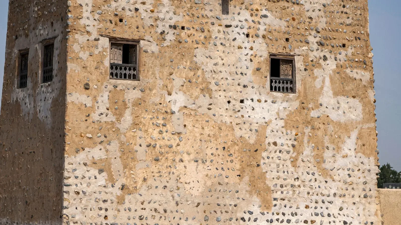 Weathered fort wall made of rounded grey and beige stones embedded in pale mud plaster, featuring three small recessed windows with dark carved wooden lattice screens, under a clear blue sky.